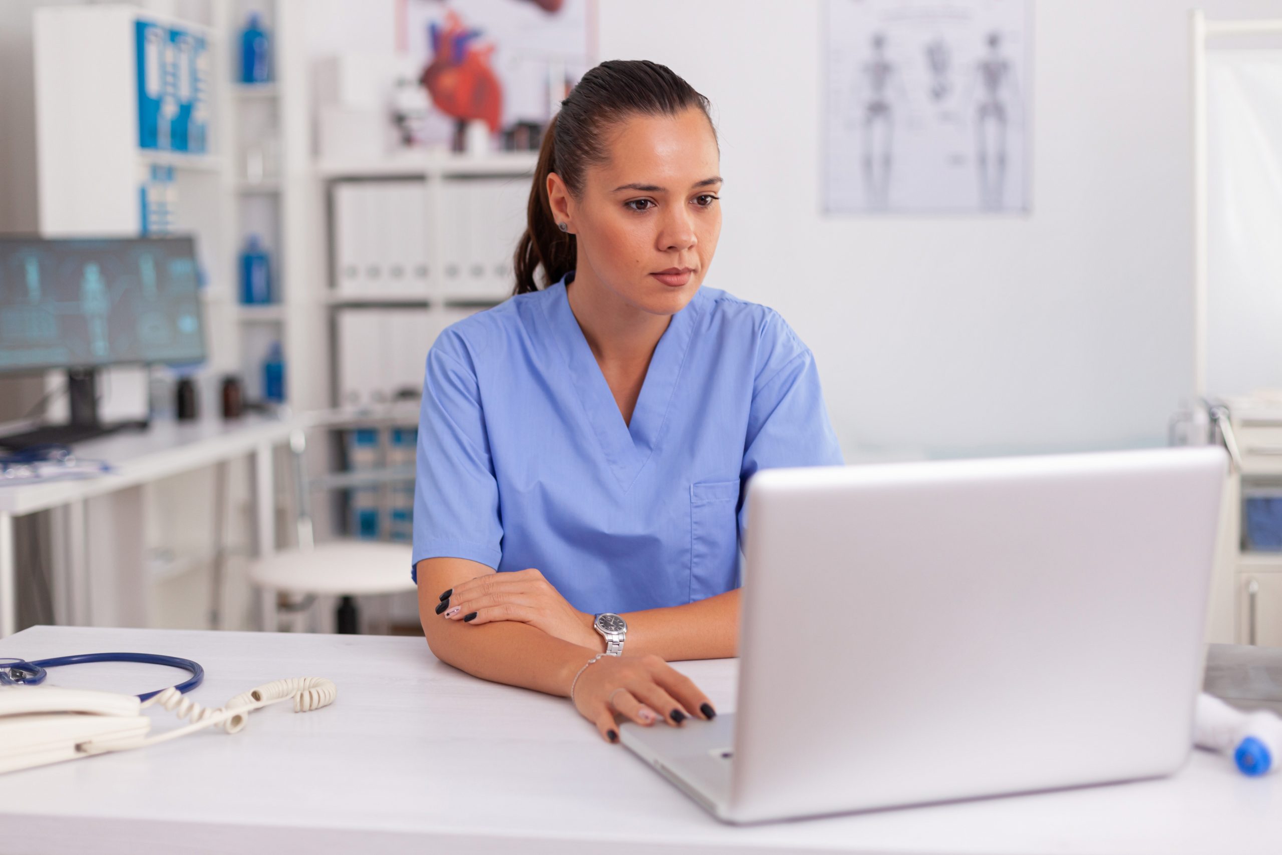 Enfermeira médica trabalhando no laptop no escritório do hospital, vestindo uniforme azul. Profissional de saúde sentado na mesa usando o computador na clínica moderna, olhando para o monitor, medicina.